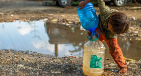 child pouring dirty water from a puddle into a larger jug